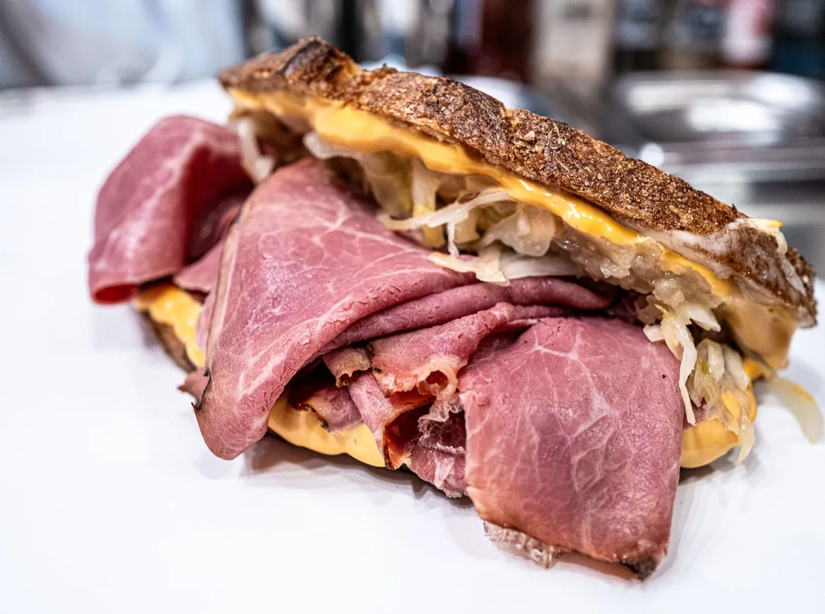 Opékaný sendvič navršený tenkými plátky růžového pastrami, rozpuštěným sýrem a kysaným zelím